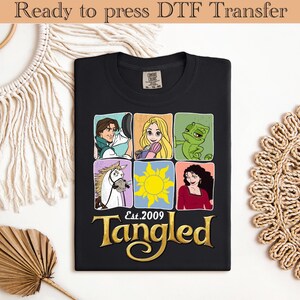 Könnte beinhalten: Schwarzes T-Shirt mit einer farbenfrohen Grafik von Charakteren aus dem Film "Rapunzel - Neu verföhnt". Das Design enthält Bilder von Rapunzel, Flynn Rider, Pascal, Maximus und der Sonne, mit dem Text "Est. 2009" und "Tangled" in Goldbuchstaben. Das Shirt ist bereit für den DTF-Transfer.