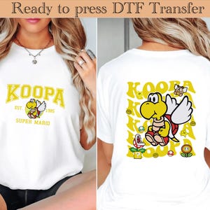 May include: White t-shirt featuring a yellow and black Koopa Troopa graphic from Super Mario. The front of the shirt displays the word "KOOPA" in yellow block letters, with the character and the text "EST. 1985 SUPER MARIO". The back has a larger Koopa graphic.
