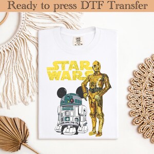 Peut inclure: T-shirt blanc avec un motif jaune "STAR WARS" et des illustrations de C-3PO et R2-D2. Le t-shirt a un col rond et des manches courtes. Le texte "Ready to press DTF Transfer" est en haut de l'image.