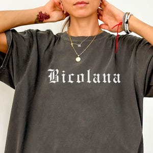 May include: A dark gray t-shirt with the word "Bicolana" printed in white, featuring a vintage, slightly faded look. The shirt has short sleeves and a relaxed fit. The word "Pepper" is visible at the bottom left corner.