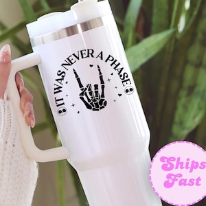May include: White insulated tumbler with a handle and a black graphic that says "IT WAS NEVER A PHASE" with a skeleton hand making a rock and roll sign. The tumbler has a white lid and a silver straw.