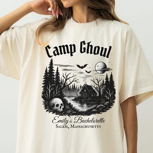 May include: Ivory oversized t-shirt with a black graphic print of a cabin in the woods, a skull, bats, and a full moon. The text "Camp Ghoul" is printed in a gothic font above the graphic. The text "Emily's Bachelorette Salem, Massachusetts" is printed below the graphic.