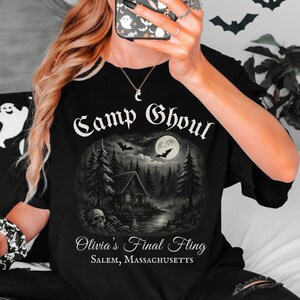 May include: Black t-shirt with a graphic of a cabin in the woods with a full moon and bats. The text "Camp Ghoul Olivia's Final Fling Salem, Massachusetts" is printed on the shirt.