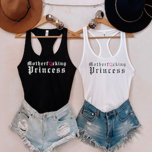 May include: Black and white racerback tank tops with the text "Motherf*cking Princess" in a gothic font. The tank tops are paired with denim shorts and are displayed with hats and sunglasses.