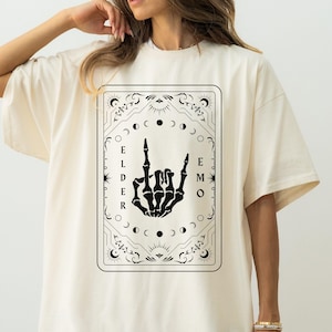May include: Ivory oversized t-shirt with a black graphic print of a skeleton hand making the rock and roll sign. The graphic is surrounded by a decorative frame with moons and stars. The text "ELDER EMO" is printed vertically on the graphic.