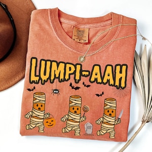 May include: A coral-coloured Comfort Colors t-shirt with the text "LUMPI-AAH" in yellow and black. The shirt features cartoon mummy characters holding a pumpkin, a lollipop, and a knife, with bats, a spider, and a tombstone graphic.