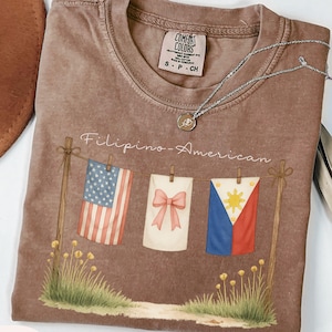 May include: Espresso-colored t-shirt with the text "Filipino-American" and a graphic of three flags: the American flag, a white banner with a pink bow, and the Philippine flag. A silver necklace and a brown hat are also visible.