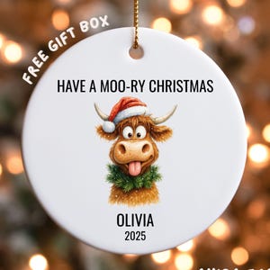 May include: White ceramic Christmas ornament with a cartoon cow wearing a Santa hat and a green wreath. The text "HAVE A MOO-RY CHRISTMAS" is above the cow, with "OLIVIA 2025" below. A gold string is attached for hanging.