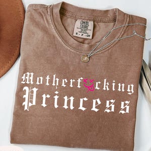 May include: Espresso-colored t-shirt with the text "Motherf*cking Princess" in white gothic font. A silver necklace with a small pendant rests on the shirt. The shirt has a crew neck and short sleeves. The shirt is a Comfort Colors brand.