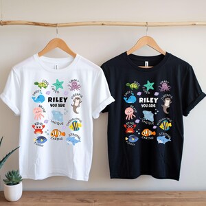 May include: Two t-shirts, one white and one black, each with a colorful sea creature design. The design includes the text "RILEY YOU ARE" surrounded by words like "sweet", "brave", and "unique".