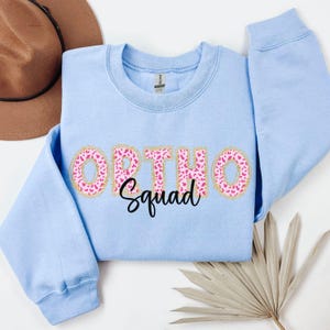 Op de afbeelding: Lichtblauwe trui met de woorden "Ortho Squad" in roze en gouden letters met een luipaardprint.