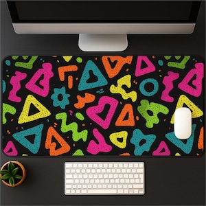May include: Black rectangular desk mat with a vibrant pattern of colorful shapes including triangles, circles, and squiggles in neon pink, orange, yellow, green, and teal. A white computer mouse sits on the mat, with a keyboard and monitor in the background.
