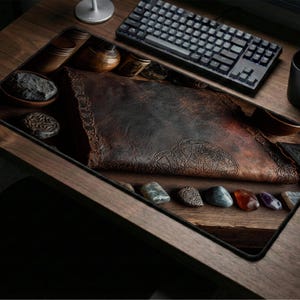 May include: A dark brown desk mat with a leather-like texture, featuring a decorative border and a row of polished stones. A black keyboard, mouse, and mug are also on the wooden desk. The mat measures approximately 76 cm long.