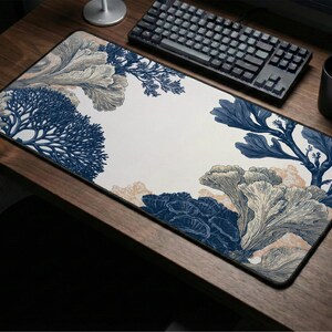 May include: Rectangular desk mouse pad with a botanical design. The pad features a white background with illustrations of blue and beige coral and plant life. A black keyboard, mouse, and mug are on the wooden desk.