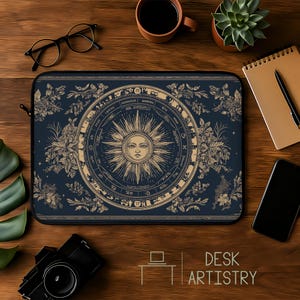 Op de afbeelding: Een marineblauwe laptoptas met een zonontwerp in het midden, omringd door dierenriemtekens en bloemmotieven. De tekst "DESK ARTISTRY" staat onderaan. De tas ligt op een houten bureau.