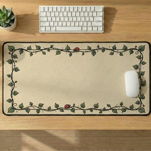 May include: A rectangular desk mat with a cream-colored surface featuring a black-lined border of winding green vines and small red ladybugs. The mat has a black stitched edge and is positioned on a wooden desk with a white keyboard and mouse.
