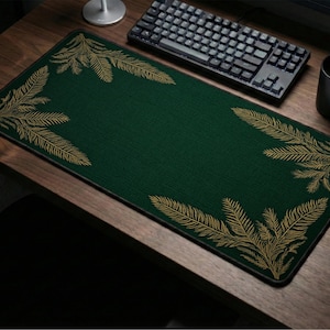 Puede incluir: Alfombrilla de escritorio rectangular verde oscuro con un diseño de hojas de helecho doradas en cada esquina. La alfombrilla está sobre un escritorio de madera, con un teclado, un ratón y una taza negros visibles.