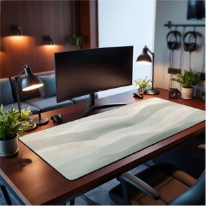 May include: A large rectangular desk mat with a wavy, watercolour-style design in shades of green and white. The mat sits on a dark wood desk, with a computer monitor, desk lamps, and plants in the background. The mat is designed for a computer setup.