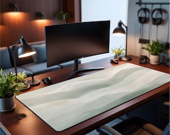 May include: A large rectangular desk mat featuring a soft, abstract watercolor pattern in shades of sage green, cream, and white. The mat has a smooth surface with a black stitched edge, positioned on a dark wood desk under a computer monitor.