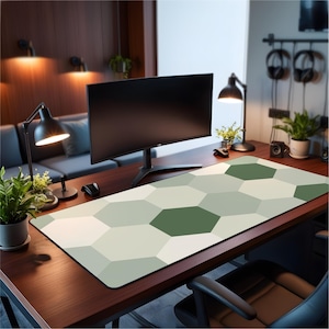 Op de afbeelding: Een rechthoekige bureaumat met een geometrisch patroon van groene, grijze en witte zeshoeken. De mat ligt op een houten bureau, met een computermonitor, bureaulamp en planten op de achtergrond.