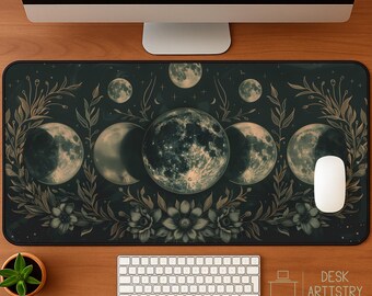 Peut inclure: Un tapis de bureau rectangulaire au fond sombre orné d'une pleine lune centrale, entourée de phases lunaires et d'illustrations botaniques complexes de feuilles et de fleurs dans des tons dorés et verts. Une souris blanche est posée à droite.