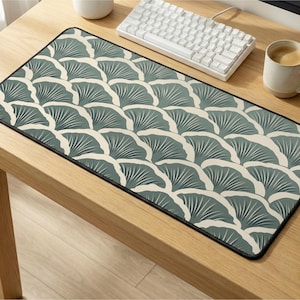 May include: A rectangular desk mat with a repeating fan-shaped pattern in teal and off-white. The mat has a black border and is placed on a light wood desk. A white keyboard and a mug of coffee are also on the desk.