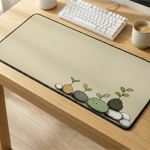 Pode incluir: Tapete de rato retangular bege com uma borda preta. A borda inferior apresenta uma ilustração caprichosa de seixos brotando em tons de cinza, verde e branco. Um teclado e uma xícara de café são visíveis no fundo.
