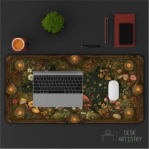 May include: A rectangular desk mat featuring a detailed forest floor illustration with mushrooms, flowers, and foliage, topped with a silver laptop and a white computer mouse. A coffee mug, a notebook, and pens are placed nearby on the desk.
