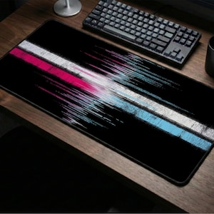 May include: Black rectangular mouse mat with a colourful abstract design. The design features a white, pink, and blue streak across the centre. A black keyboard, mouse, and mug are on a wooden desk.