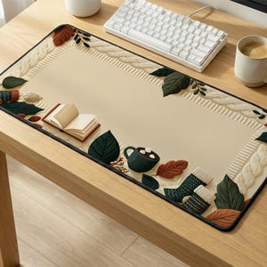Puede incluir: Una alfombrilla de escritorio rectangular con un centro color crema y un borde con patrón de punto trenzado, hojas verde oscuro, hojas marrones, un pequeño libro abierto, una taza verde oscuro con malvaviscos y calcetines verdes estampados.