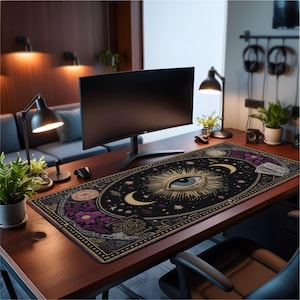 Puede incluir: Una alfombrilla de escritorio grande, negra y dorada con un diseño de ojo y celestial. La alfombrilla presenta detalles florales morados y un borde con patrones intrincados. Un monitor de ordenador, una lámpara de escritorio y un ratón están sobre el escritorio de madera.