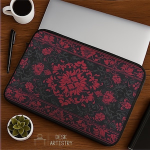 May include: A black laptop sleeve with a red floral pattern. The design features a central floral medallion and borders with repeating floral motifs. The sleeve is on a wooden surface, with a coffee cup, pen, and small plant nearby.