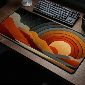 May include: A rectangular mouse mat with an abstract landscape design. It features layered orange, yellow, and grey shapes, reminiscent of a sunset or mountain range. The mat is on a wooden desk, with a keyboard, mouse, and mug visible.