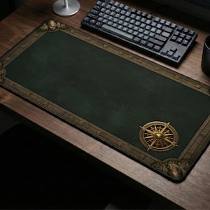 May include: A rectangular desk mat featuring a dark green center and a decorative border with gold-toned corner accents. A gold compass rose graphic is positioned in the lower right corner of the mat, which is set on a wooden surface.