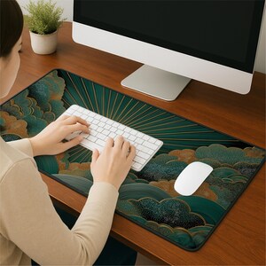 May include: A rectangular desk mat featuring a dark teal and gold pattern of stylized clouds and radiating sunbeams. The mat is positioned on a wooden desk surface beneath a white computer keyboard and a white computer mouse.