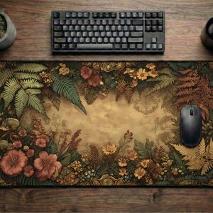 Op de afbeelding: Een rechthoekige muismat met een gedetailleerd botanisch ontwerp met varens, bloemen en bladeren in groen, bruin en oranje tinten. Een zwarte computermuis en toetsenbord liggen op een houten oppervlak.