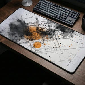May include: A rectangular white mouse mat with a black border, featuring an abstract design with black, orange, and grey paint splatters and lines. A black keyboard, mouse, and mug are on a wooden desk.