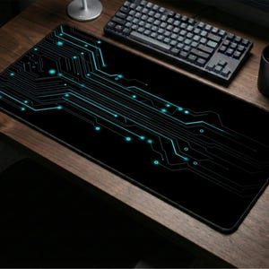 Op de afbeelding: Een zwarte rechthoekige muismat met een turquoise print van een printplaat. De muismat ligt op een houten bureau, met een zwart toetsenbord, muis en mok. Het printplaatontwerp heeft lijnen en stippen.