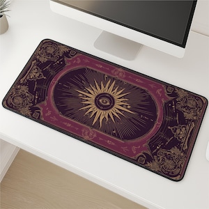 Puede incluir: Alfombrilla de escritorio rectangular con un fondo morado oscuro y un diseño de sol dorado con un ojo en el centro. La alfombrilla tiene un borde negro y detalles dorados ornamentados en los bordes, creando una estética mística.