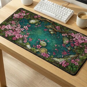 May include: A rectangular desk mat featuring a detailed illustration of a tranquil pond surrounded by blooming pink cherry blossom trees, green foliage, and scattered stones, with fallen pink petals floating on the water surface.