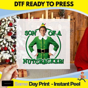 May include: A DTF (Direct to Film) ready-to-press transfer featuring a green and white graphic of a character from the movie "Elf" with the text "SON OF A NUTCRACKER!". The image is held in front of a Christmas tree and fireplace.
