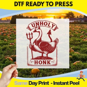 May include: A DTF (Direct to Film) ready to press transfer featuring a red illustration of a goose with a pitchfork and devil tail, with the text "UNHOLY HONK". The background shows a pumpkin field.