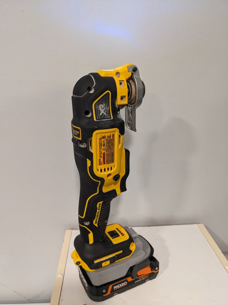 May include: A yellow and black cordless oscillating tool with a brushless motor. The tool is attached to a black and orange battery pack with the word "RIDGID" printed on it.