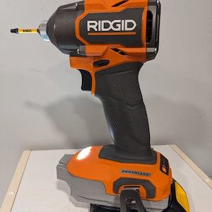 May include: An orange and black cordless drill with a black battery pack. The drill has a black handle and a silver trigger. The battery pack is labelled "DEWALT 20V MAX* LITHIUM ION". The drill is labelled "RIDGID" and "BRUSHLESS".