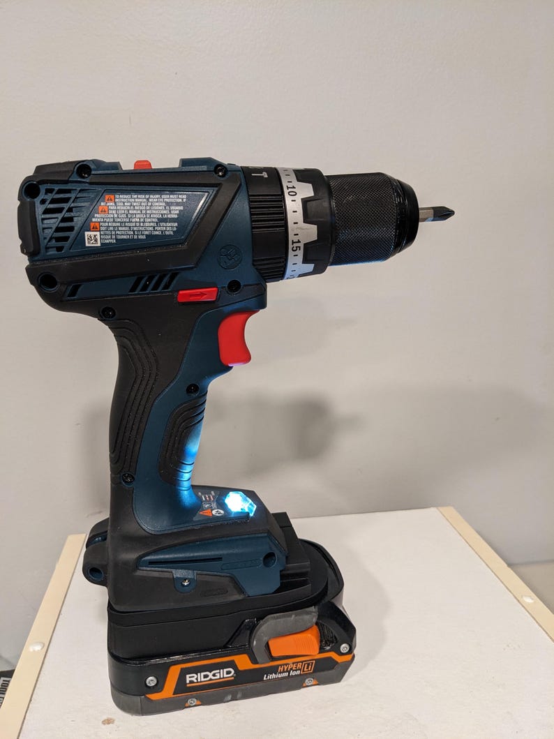 Puede incluir: Un taladro inal&aacute;mbrico azul y negro con un portabrocas negro y un dial de ajuste de torque plateado. El taladro est&aacute; conectado a una bater&iacute;a naranja y negra que dice "RIDGID HYPER LI Lithium Ion".