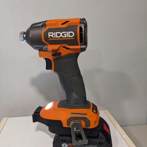 Op de afbeelding: Een oranje en zwarte draadloze schroevendraaier met een ingebouwde LED-lamp. De schroevendraaier is gemarkeerd met "RIDGID" en "BRUSHLESS".