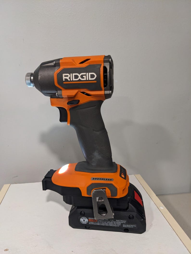 Op de afbeelding: Een oranje en zwarte draadloze schroevendraaier met een ingebouwde LED-lamp. De schroevendraaier is gemarkeerd met "RIDGID" en "BRUSHLESS".