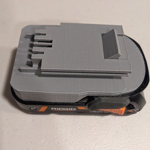 Pu&ograve; includere: Un supporto per batteria grigio stampato in 3D per uno strumento elettrico Ridgid. Il supporto ha uno slot per la batteria e un pulsante di rilascio.