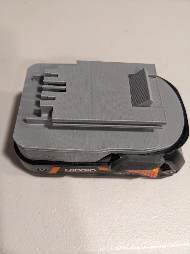 Pu&ograve; includere: Un supporto per batteria grigio stampato in 3D per uno strumento elettrico Ridgid. Il supporto ha uno slot per la batteria e un pulsante di rilascio.