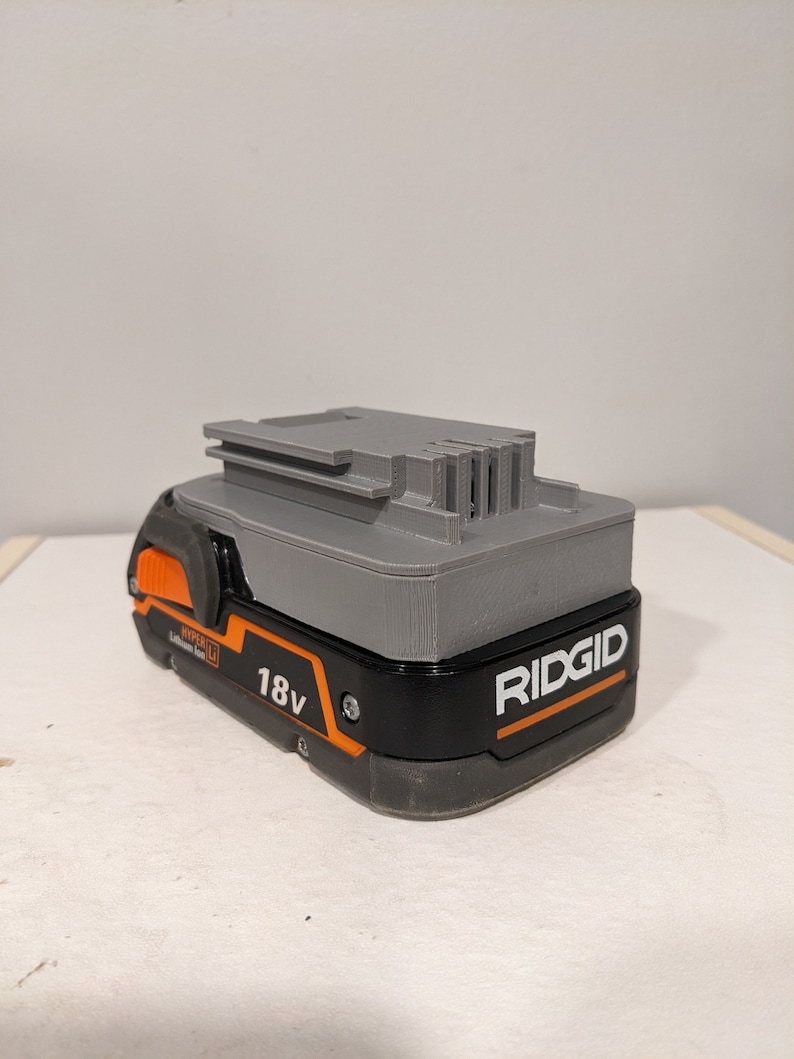 Pu&ograve; includere: Una batteria Ridgid da 18 V di colore grigio e nero con un pulsante arancione e il testo "HYPER LI Lithium Ion" e "RIDGID" sul lato.